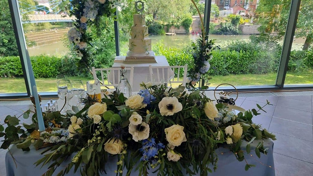 Reception table of soft cream and blue flowers, low enough for conversation, with taper candles and a gold hoop behind the cake